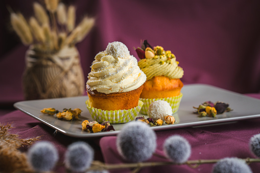 Side view of two gluten free carrot cake muffins topped with cream, displayed on a chic white plate and sprinkled with crunchy pistachios. All arranged on a purple studio surface fabric and decorated with dried plants. Side view, full length image.