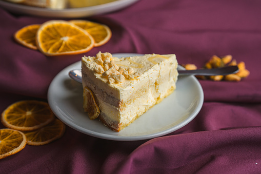 A studio shot of a gluten free, professionally made slice of cake arranged with dried orange slices, all displayed on purple fabric. Full length image, high angle view.