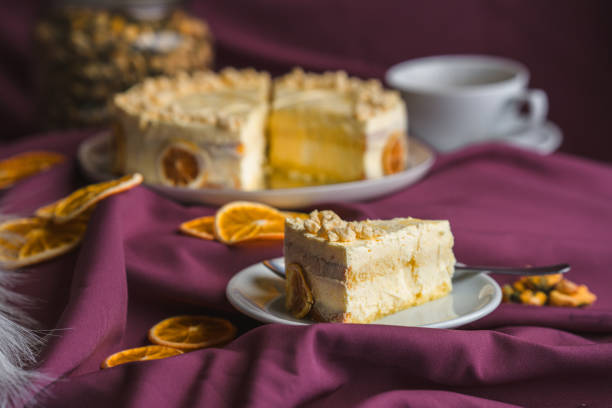 A studio shot of a round, professionally made cake arranged with dried orange slices, a cup of tea,  nuts in a jar in the background, focus on the slice on a separate ceramic plate, all arranged on purple fabric. Full length image.