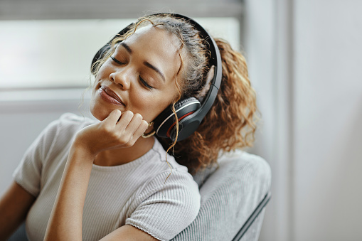 Music, relax and woman on a chair for peace, calm and happiness in the living room of her house. Face of a young girl sleeping with audio, radio or podcast in her headphones in the lounge of her home