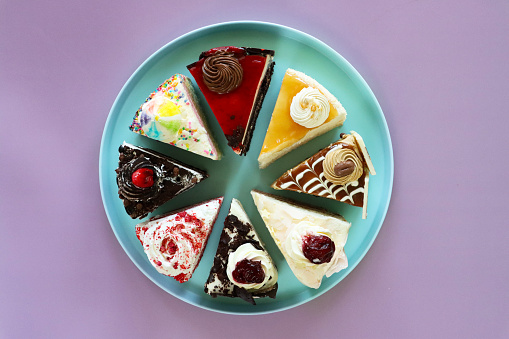 Stock photo showing elevated view of slices of angel food cake, chocolate and cherry cake, Black Forest gateau, red velvet, rainbow cake, coffee and marbled chocolate gateau and salted caramel cake displayed on a pastel blue plate.