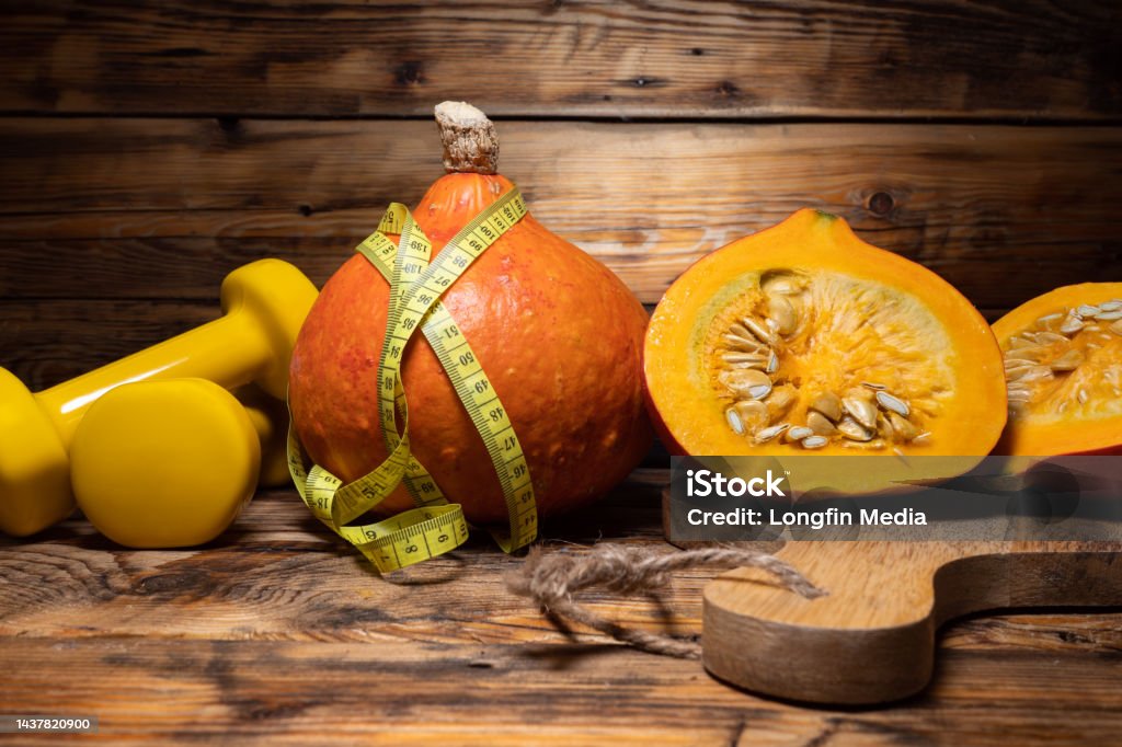 Gym dumbbells with Hokkaido pumpkins and tape measure. Autumn or fall rustic composition. Hokkaido pumpkins or red kuri squash with tape measure or measuring tape and dumbbells. Healthy fitness lifestyle autumn rustic composition for Halloween or Thanksgiving. Gym workout and sport training fall concept. Thanksgiving - Holiday Stock Photo