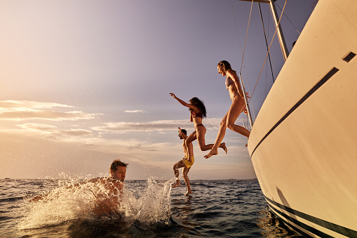 Group of playful friends having fun while jumping into sea from a yacht during summer sunset. Copy space.