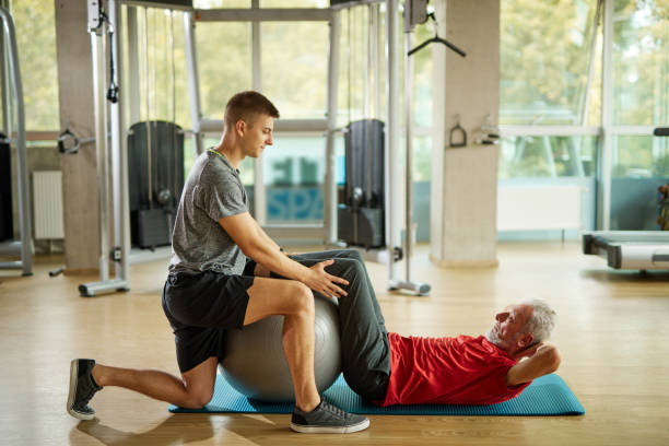 Personal trainer helping senior man exercising with fitness ball at the gym stock photo