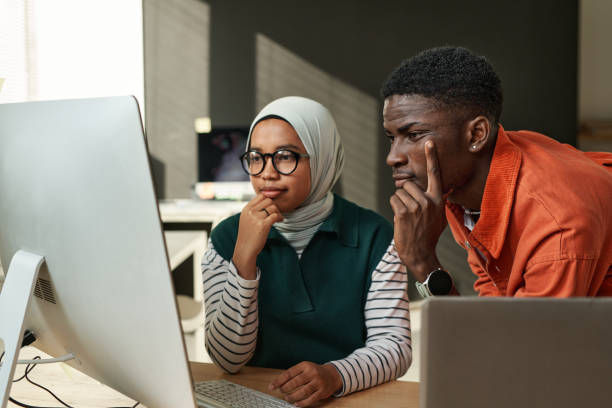 deux jeunes ingénieurs informatiques interculturels sérieux regardant un écran d’ordinateur - emigration and immigration photos et images de collection