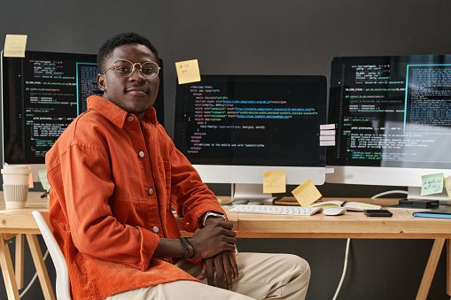 Young IT support manager looking at camera while sitting by workplace in front of computer monitors with decoded data on screens
