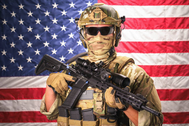 istock Armed special forces soldier hold machine gun dressed in ammunition posing on USA flag on background. Army soldier in camouflage clothing. Flag of United States of America on background 1437828247