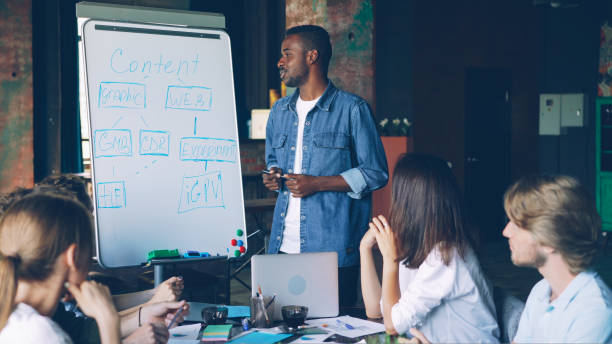 istock African American business trainer is working with whiteboard pointing at chart and speaking while company employees are learning and talking sitting at table in office. 1437829480