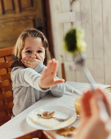 Small girl covering her mouth while refusing to eat a broccoli in dining room.