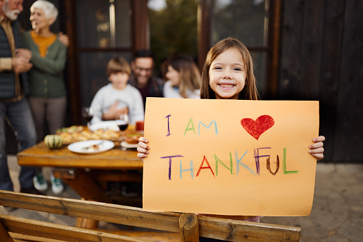 Happy girl holding 'I'm thankful' sign and looking at camera during a meal with her family on a patio.