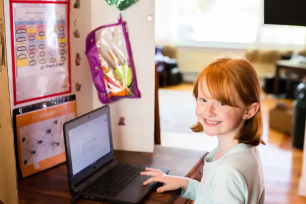 Young girl using laptop. Homeschooling, hand on keyboard at kitchen table with divider around her.