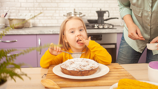 Funny little girl child tasting sweet powdered sugar, decorating cake, having fun and smiling, cooking in kitchen. looks into camera