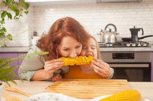 child eats sweet boiled corn in purple kitchen of house. Funny girl at table. mother and daughter eat together