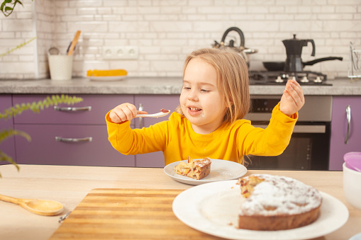 funny little girl eats plum pie with sugar, happy and satisfied, holding spoon on own. Laughs