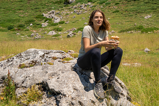 Young caucasian woman sitting on a rock prepared to eat a sandwich in the nature after a trekking.
