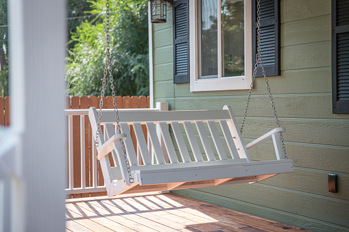 A white bench swing hangs over a home porch decking on a sunny afternoon.