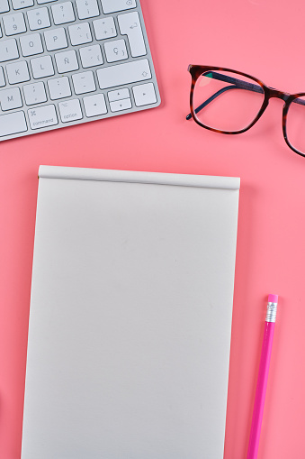 A vertical top view of glasses, a notepad, a pencil, and a keyboard on a pink background