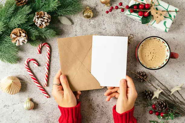 Photo of Desktop of woman hands in red sweater holding blank paper and envelope . Flat lay of gray background with cup of coffee and Christmas decoration. Top view mock up and copy space for text