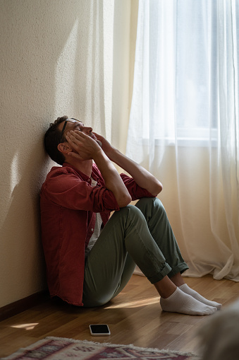 Upset depressed young man sitting with closed eyes on floor at home, getting unexpected bad news over phone. Frustrated guy suffering from relationship break up, cannot forget about ex-girlfriend