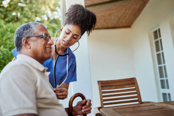 Healthcare, disability and an elderly man and nurse on retirement or nursing home patio. Senior care, happy  grandpa with cane and black woman caregiver checking heart outside in garden at hospice stock photo
