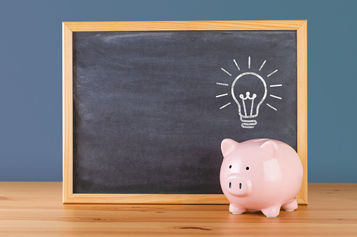 Front view of a piggy bank in front of a blackboard with a drawing light bulb on top of the piggy bank. There is a useful copy space at the left side of the blackboard. Objects are on a wooden desk against a blue wall.