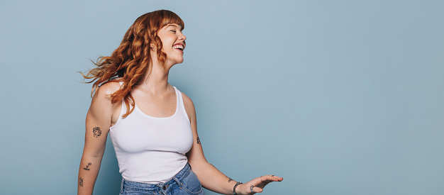 Smiling woman with ginger hair dancing and having fun against a blue background. Woman with arm tattoos celebrating while standing in a studio. Happy young woman wearing a tank top and jeans.