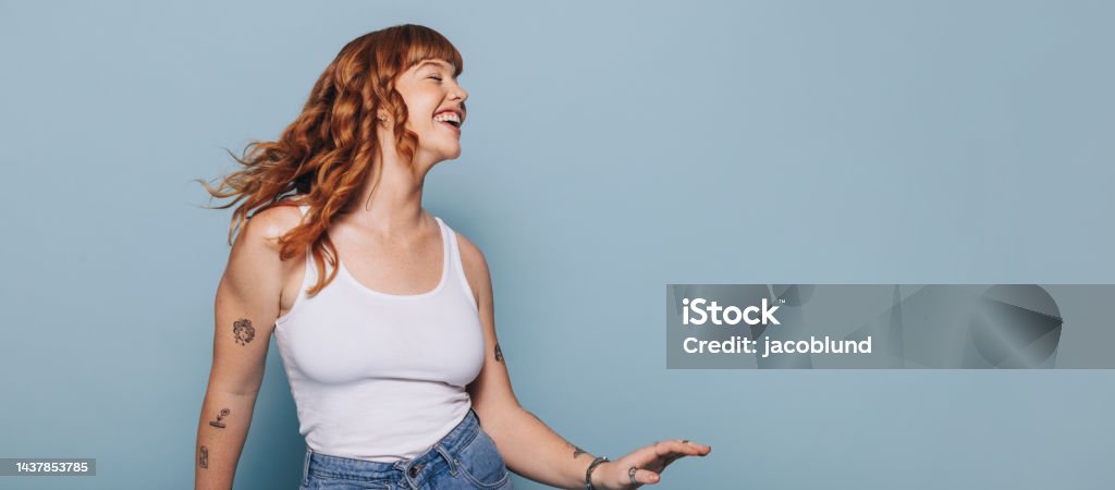 Woman with ginger hair dancing and having fun in a studio Smiling woman with ginger hair dancing and having fun against a blue background. Woman with arm tattoos celebrating while standing in a studio. Happy young woman wearing a tank top and jeans. Women Stock Photo