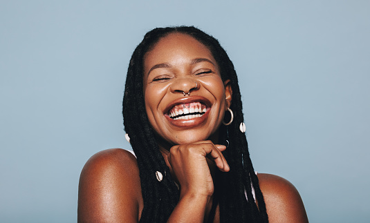 African woman with face piercings smiling cheerfully in a studio. Happy young woman wearing dreadlocks and piercing jewellery against a blue background. Female hipster feeling confident in her style.