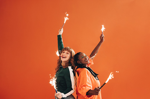 Female friends smiling at the camera while lifting bengal lights in celebration. Two vibrant young women having fun with sparkers in a studio. Two best friends making happy memories together.