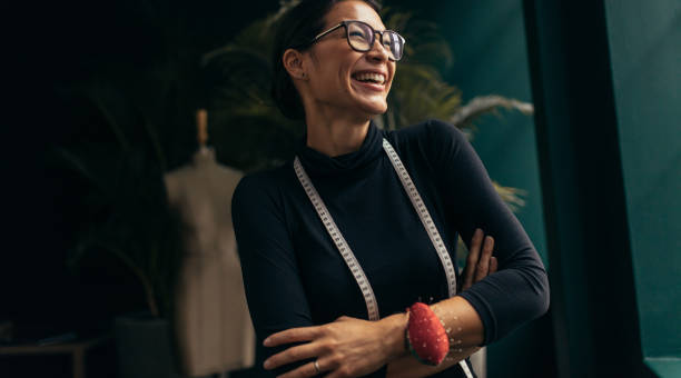 Female fashion designer laughing in her studio Laughing female fashion designer in her studio. Asian woman standing with measuring tape around neck and looking away. fashion designer stock pictures, royalty-free photos & images