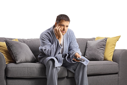 Bored young man in a bathrobe holding a remote control and sitting on a sofa isolated on white background