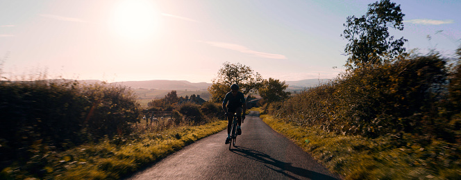 Young adult cycling on a professional road bike wearing lycra and a helmet on a small country road. He is cycling towards the camera in silhouette.
