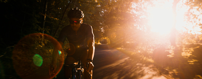 Young adult cycling on a professional road bike wearing lycra and a helmet along a country road. He is looking into the camera with the sun setting in the background.