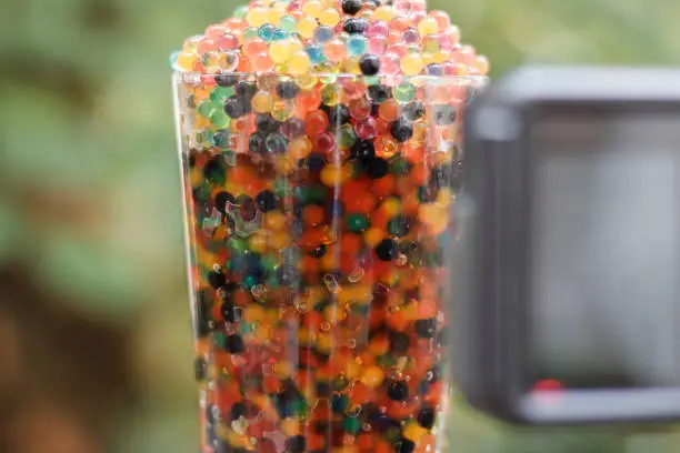 Close up on a glass filled with colored balls. An action camera set vertically on a tripod is recording the scene. Focus on the glass, camera blurred.