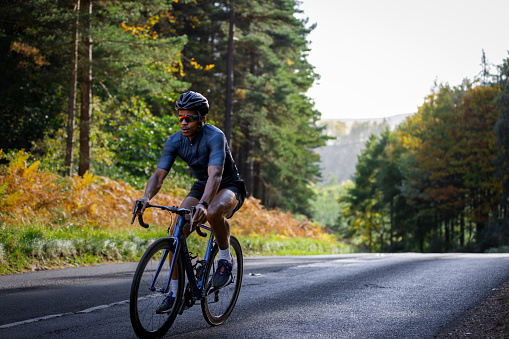 Young adult cycling on a professional road bike wearing lycra and a helmet on the road surrounded by trees. He is determined and concentrates on what he is doing.