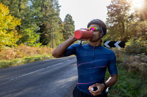 Young adult wearing lycra and a helmet on the roadside surrounded by trees. He is drinking water from his bottle, and taking a break from the bike ride he is on.