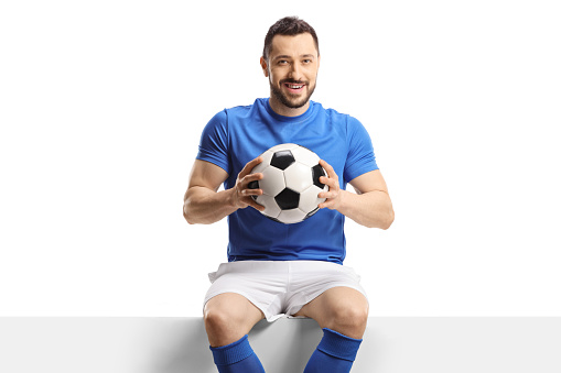 Football player sitting on a blank panel and holding a soccer ball isolated on white background