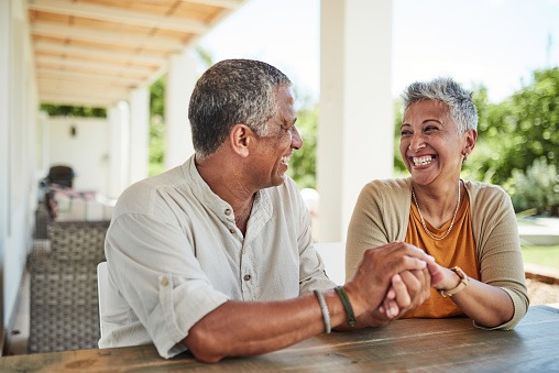 Senior couple holding hands by outdoor table in retirement home on sunny day. Bonding, love and affection of happy retired black people sitting together laughing, smiling and relax outside in summer
