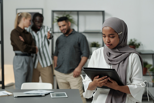 Young Muslim businesswoman in hijab holding black folder with documents against group of intercultural colleagues mocking at her