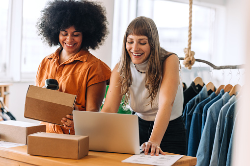 Happy businesswomen using a laptop while preparing delivery parcels in a thrift store. Two small business owners making plans for a shipment. Female entrepreneurs running an online clothing store.