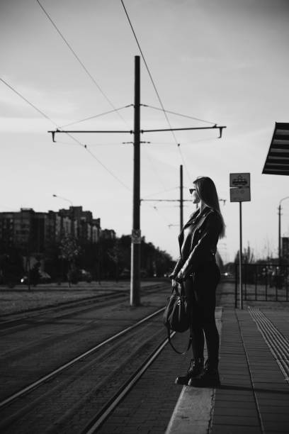 Young woman standing on the tram station and waiting for a tram. Wearing solid long sleeve bodycon one piece jumpsuit, a black leather jacket and smooth platform boots