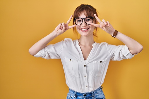 Young beautiful woman wearing casual shirt over yellow background doing peace symbol with fingers over face, smiling cheerful showing victory