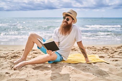 Young redhead man reading book sitting on the towel at the beach.