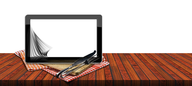 Blank digital tablet computer with white screen for recipes or food menu. On a wooden kitchen counter with a cutting board, checkered tablecloth and silver cutlery.