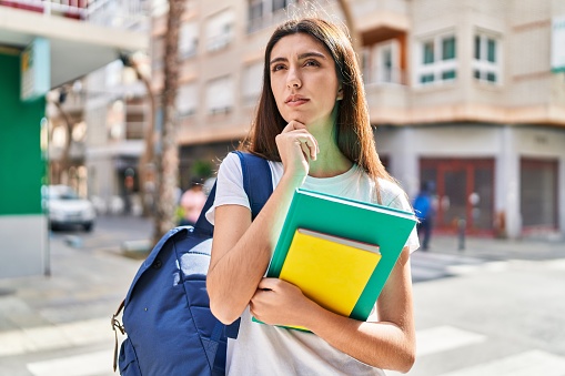 Young beautiful hispanic woman student holding books with doubt expression at street