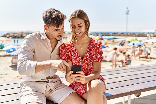 Young hispanic couple on vacation using smartphone sitting on bench at the beach.