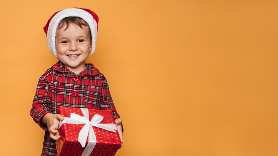 Studio photo of a baby boy in Christmas pajamas and a hat on a yellow background with a bright red gift box in his hands. A place for your text, advertising