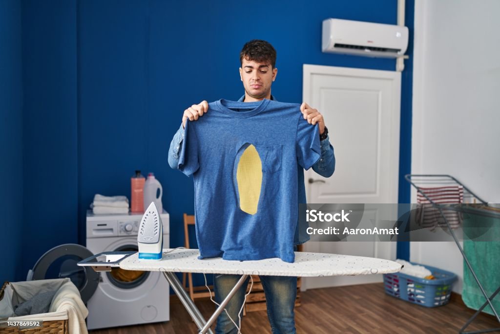 Young hispanic man ironing holding burned iron shirt at laundry room relaxed with serious expression on face. simple and natural looking at the camera. Burning Stock Photo