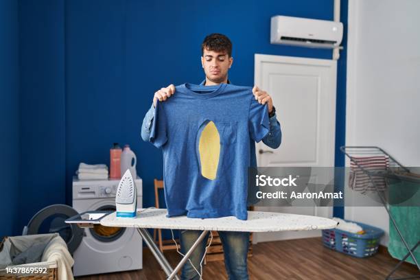 Young Hispanic Man Ironing Holding Burned Iron Shirt At Laundry Room Relaxed With Serious Expression On Face Simple And Natural Looking At The Camera Stock Photo - Download Image Now