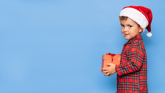 A happy little boy in Christmas pajamas and a hat with a gift box. A place for your text. Studio shot on a blue background. The concept of New Year and Christmas